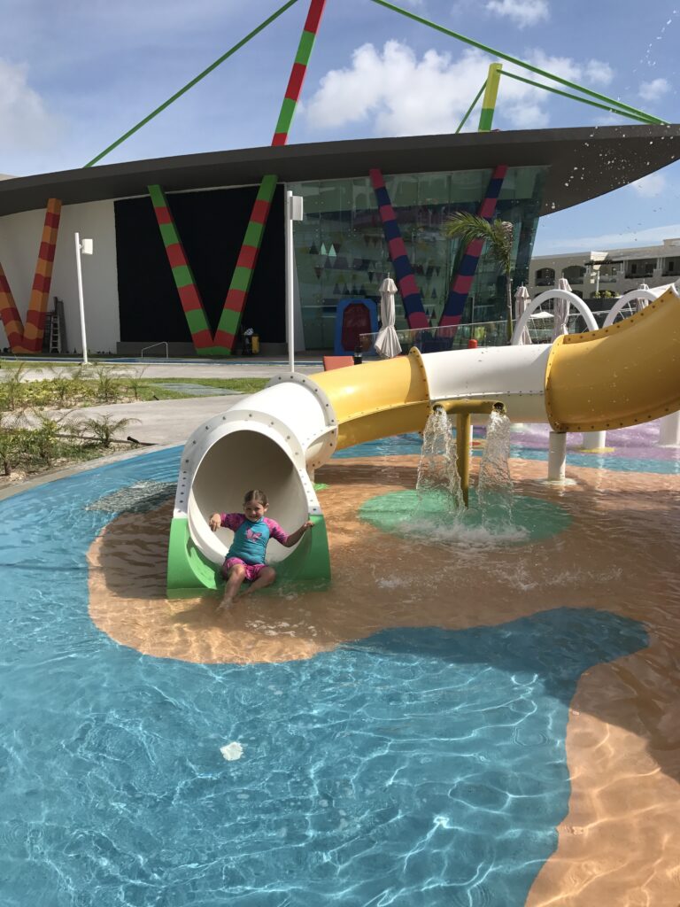 Harper, wearing a pink and turquoise swimsuit, slides down a water slide into a shallow splash pool at the Grand Moon Palace resort, with a colorful kids’ club building in the background.