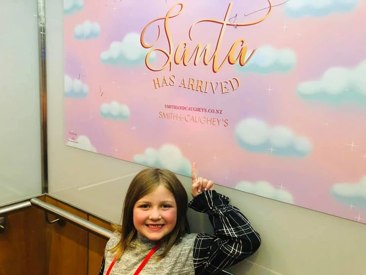 Harper smiling and pointing to a pastel sign reading “Santa Has Arrived” at Smith & Caughey’s in Auckland, New Zealand.
