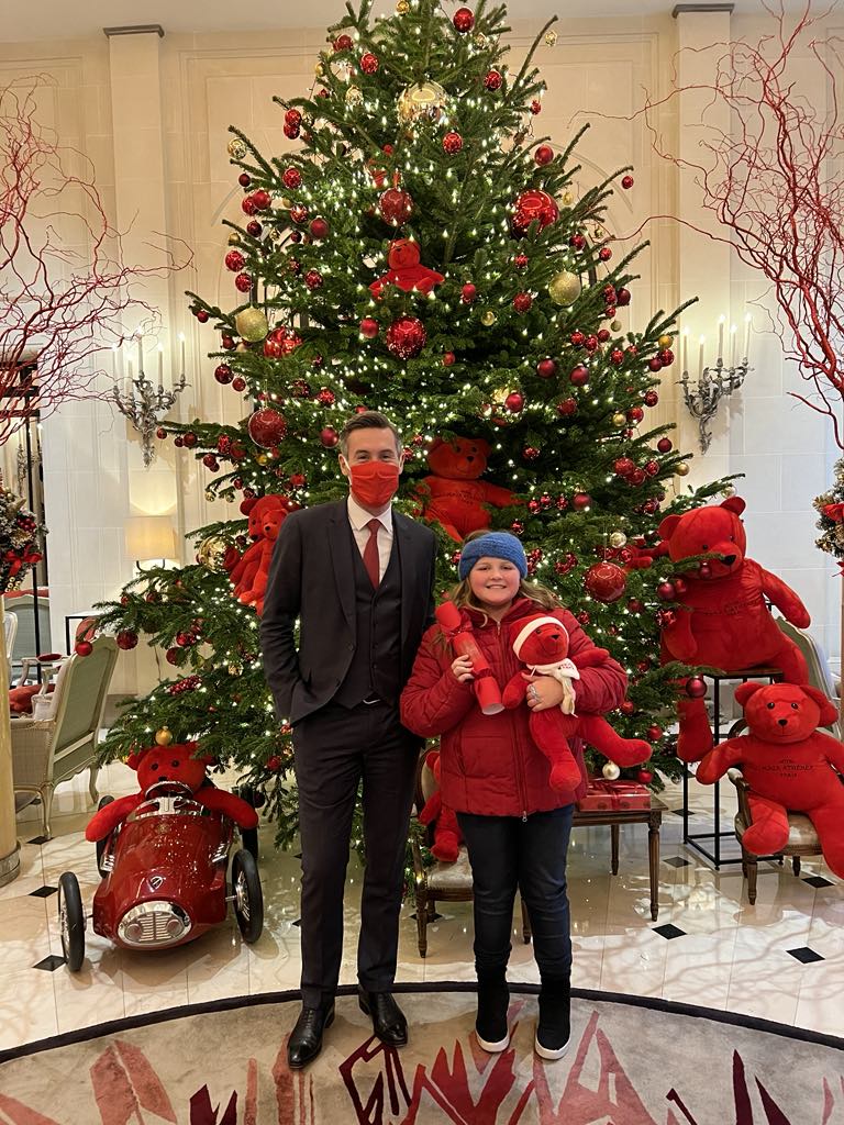 Kid traveler Harper standing with the general manager of a luxury hotel in front of a beautifully decorated Christmas tree, smiling while holding a festive red teddy bear gifted during a memorable holiday stay.
