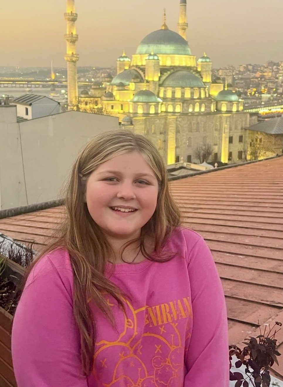 Harper smiles on a rooftop in Istanbul with the illuminated domes and minarets of a grand mosque glowing behind her at sunset.