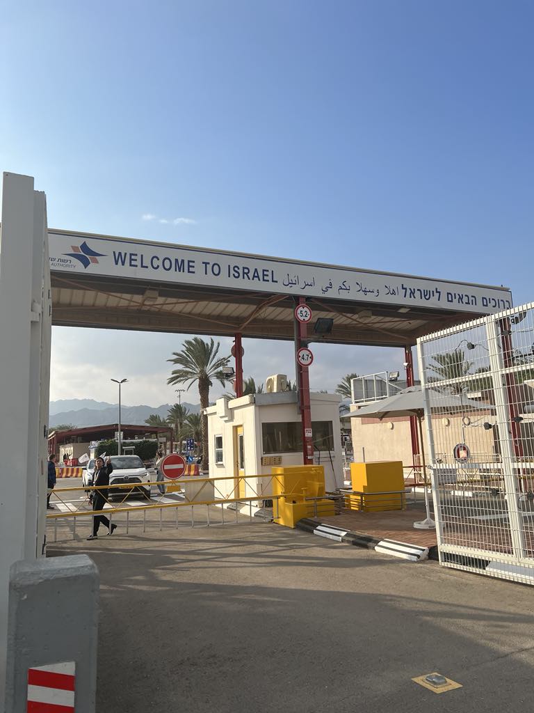 View of the Israel-Jordan border crossing at the Sheikh Hussein Bridge, with signage in Hebrew, Arabic, and English, and flags of both nations flying above.