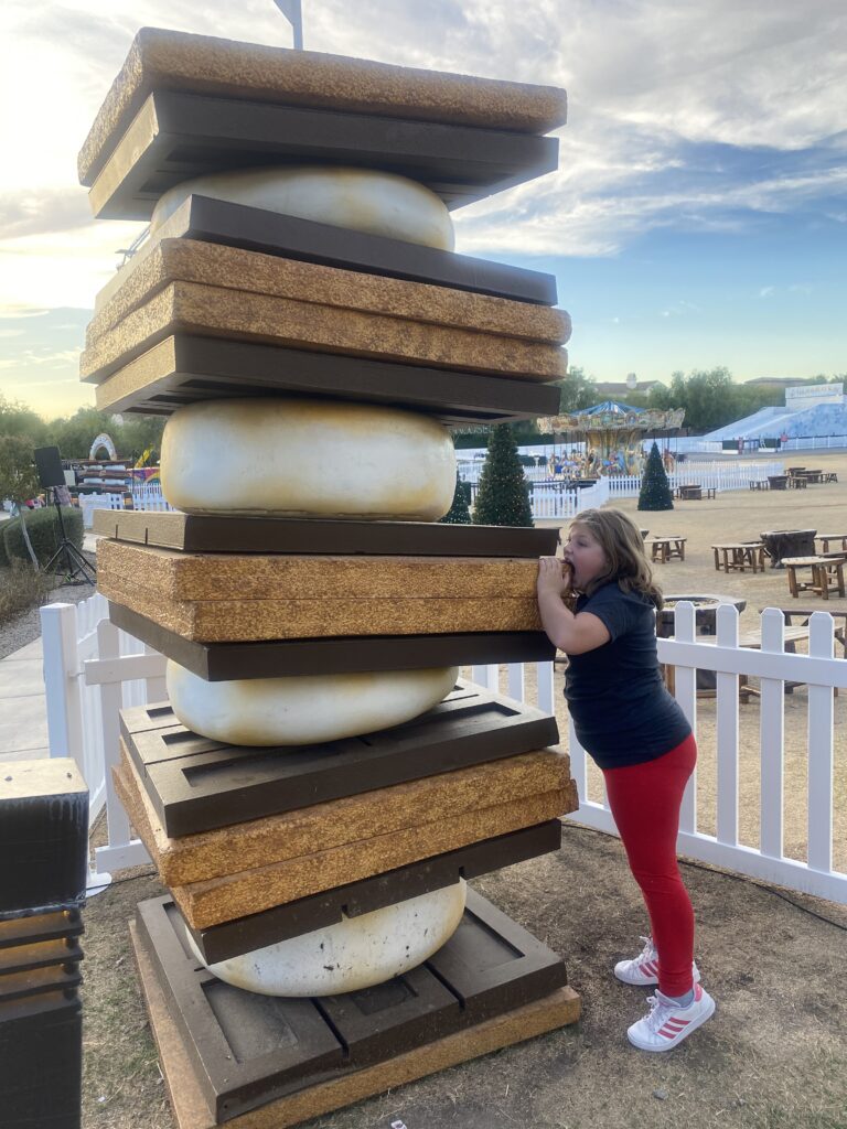 kid traveler Harper pretending to take a bite out of a giant s’more decoration at the Fairmont Scottsdale Princess during the whimsical “Christmas at the Princess” holiday celebration.